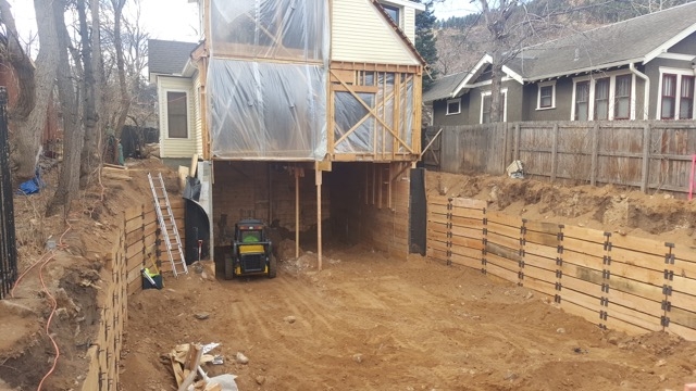 Deep residential excavation beneath a raised home with reinforced retaining walls and construction equipment.